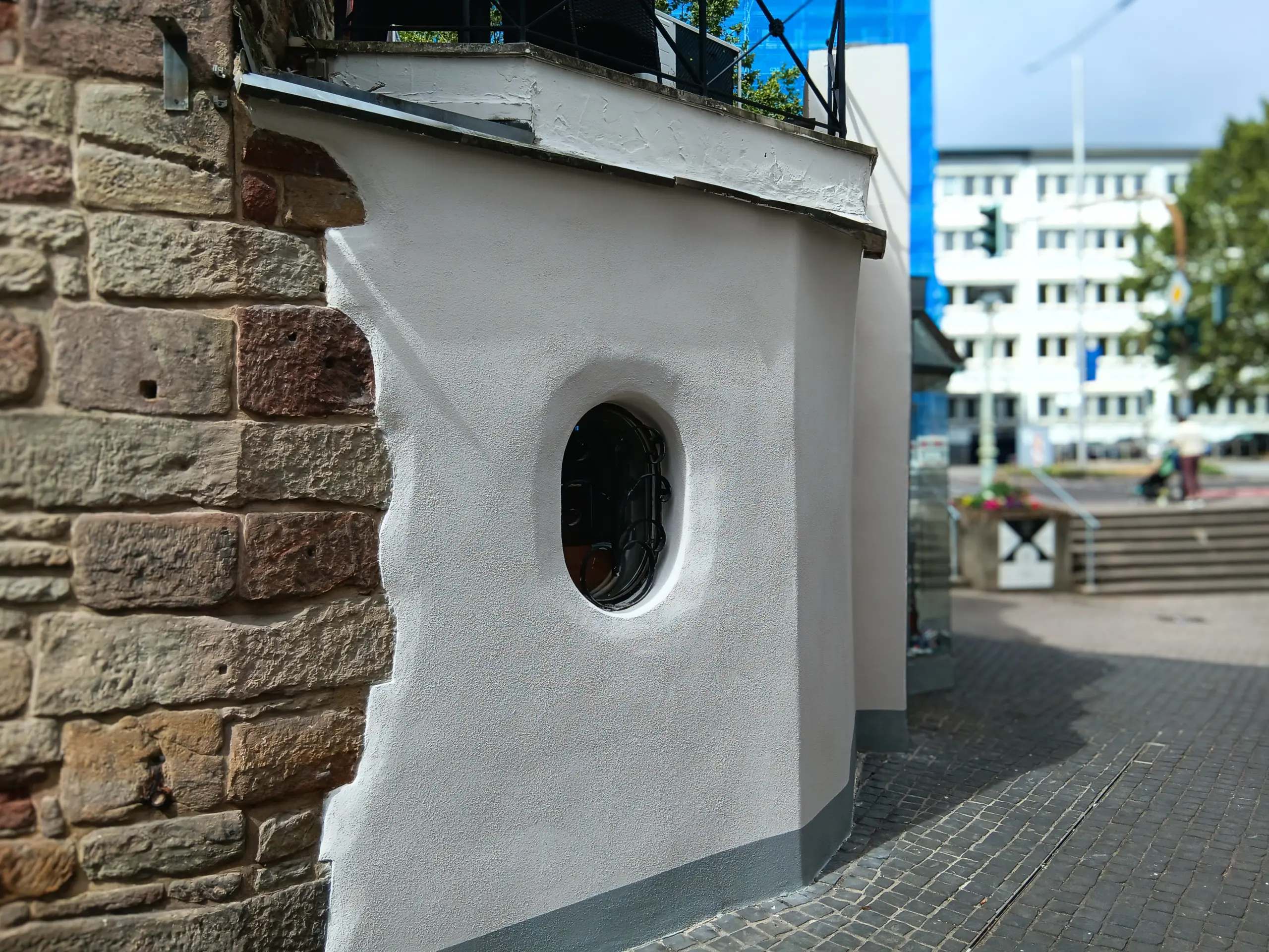 Eine teils freigelegte Sandsteinmauer, die mit frischem weißem Außenputz renoviert wurde; ein rundes, vergittertes Fenster verleiht der historischen Fassade einen besonderen Akzent.