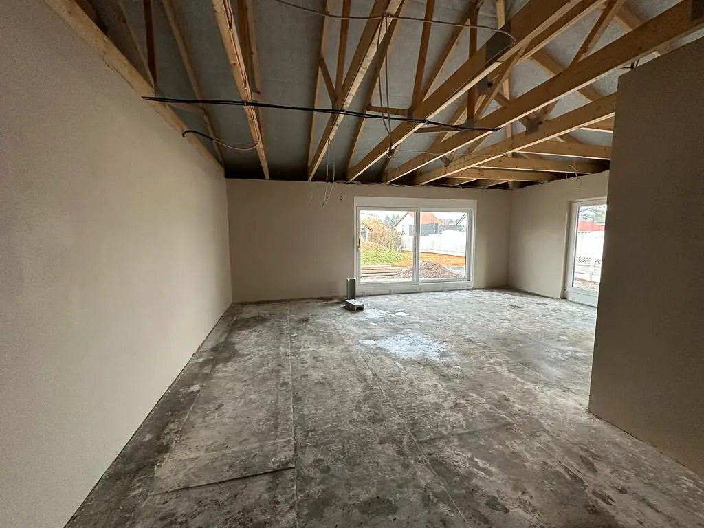 Ein großer, unfertiger Wohnraum mit verputzten Wänden, freiliegender Holzkonstruktion an der Decke und rohem Boden; durch die großen Fenster ist eine Nachbarschaft im Freien zu sehen.